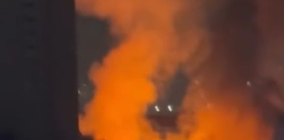 Chamas intensas e fumaça densa se elevam de um edifício, iluminando a cena com uma luz laranja brilhante