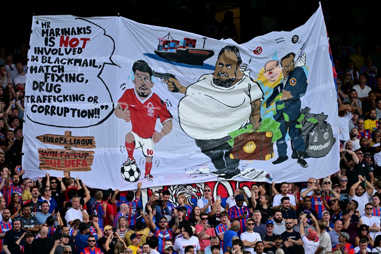 Torcida do Crystal Palace exibe faixa em que John Textor aparece com um nariz de palhaço (No canto direito, 'no colo' de um dirigente da Uefa vestido de azul)