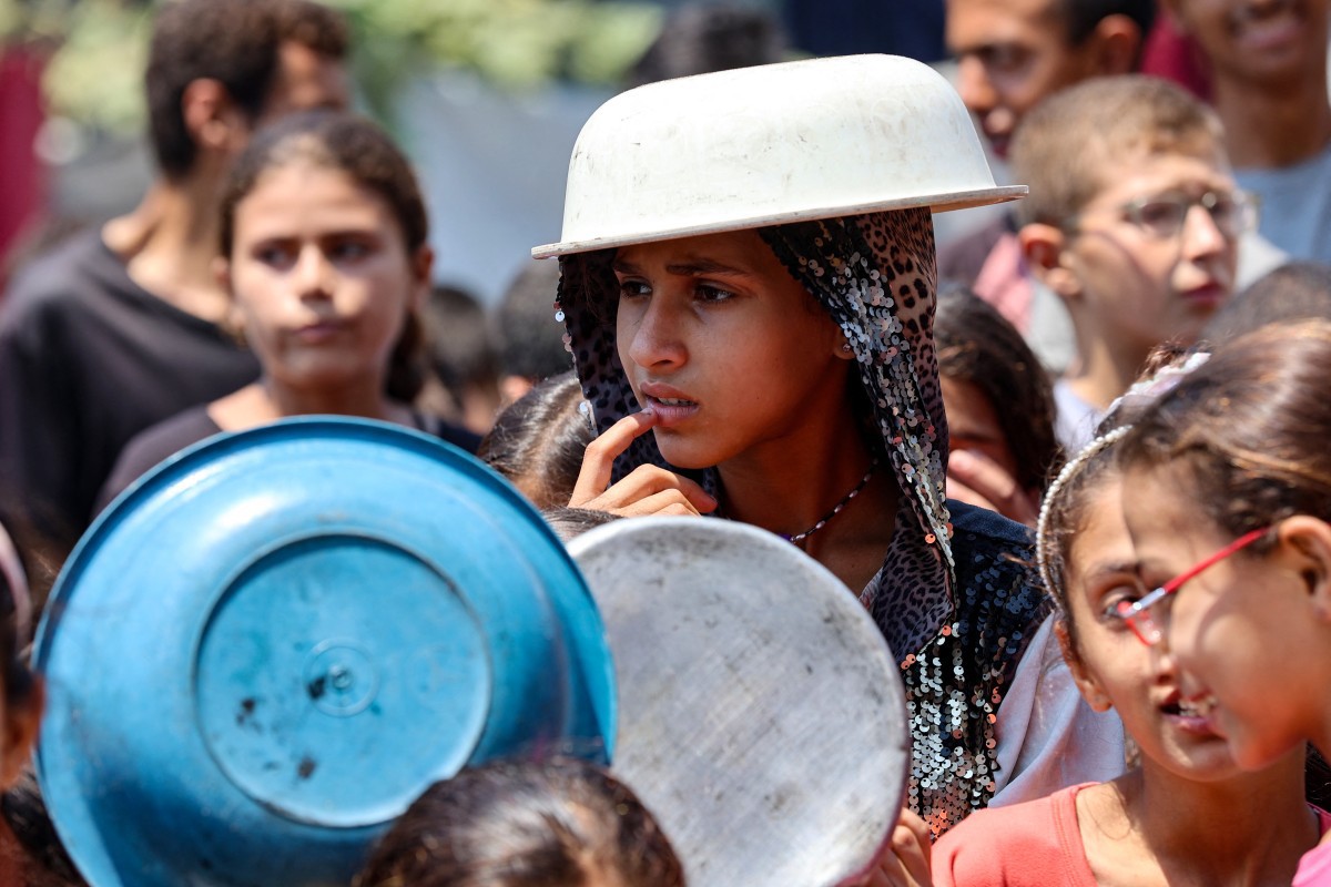 Menina palestina cobre a cabeça com uma panela para se proteger do sol, enquanto espera em um ponto de distribuição de alimentos em Gaza