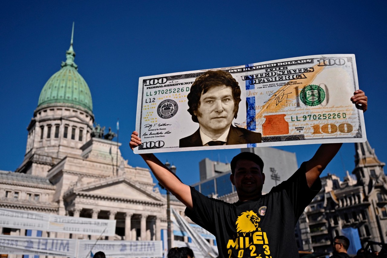 Apoiador de Javier Milei segura um cartaz em forma de nota de dólar com o rosto do presidente diante da Casa Rosada, sede do governo argentino