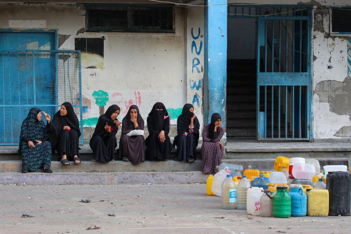 Mulheres palestinas sentam-se perto de seus galões enquanto aguardam um caminhão de distribuição de água