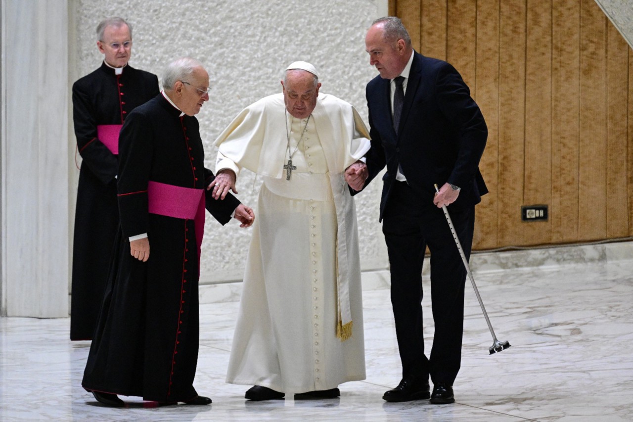 Após o tropeço, o Papa Francisco precisou ser auxiliado por dois funcionários até se encaminhar a sua cadeira no palco do auditório