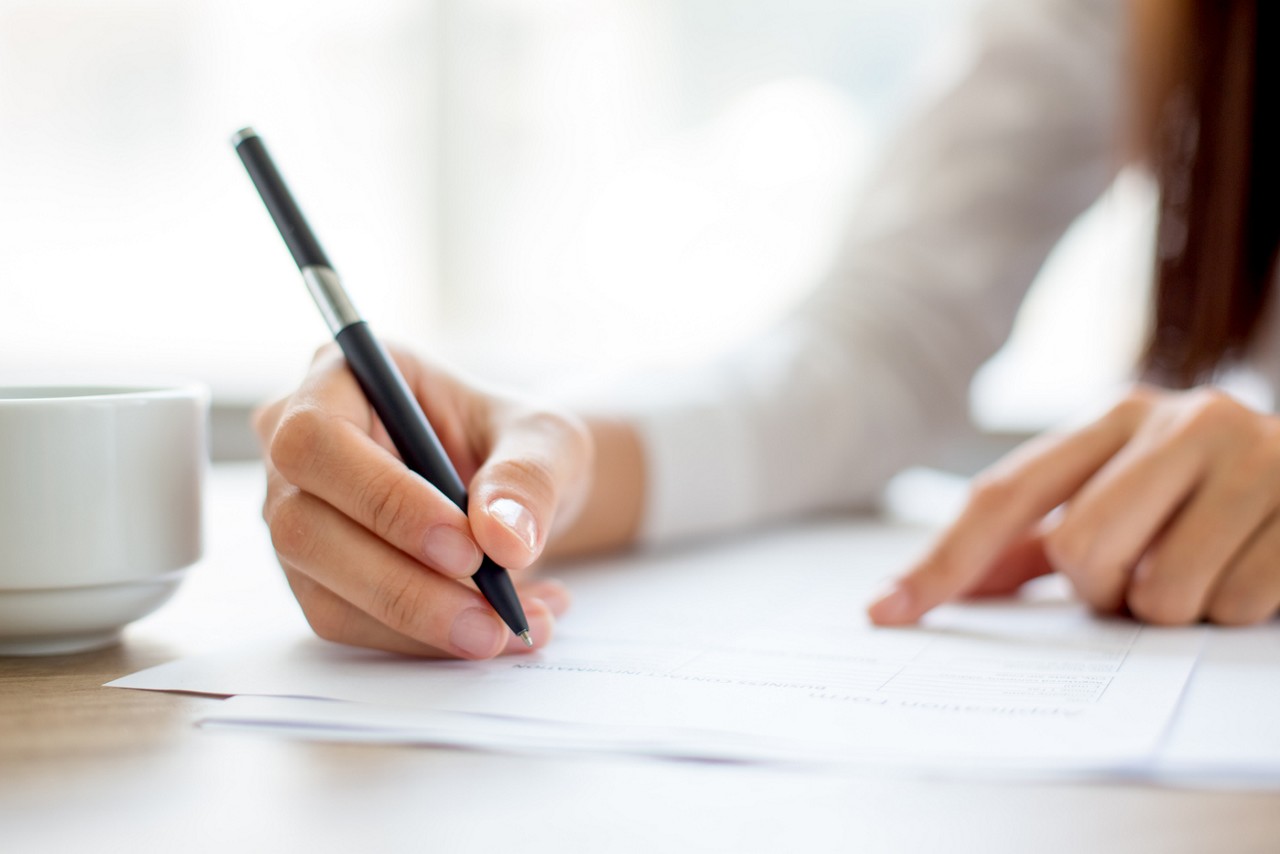 Hand of young businesswoman writing on paper or signing contract at table in office, Hand of young businesswoman writing on paper or signing contract at table in office