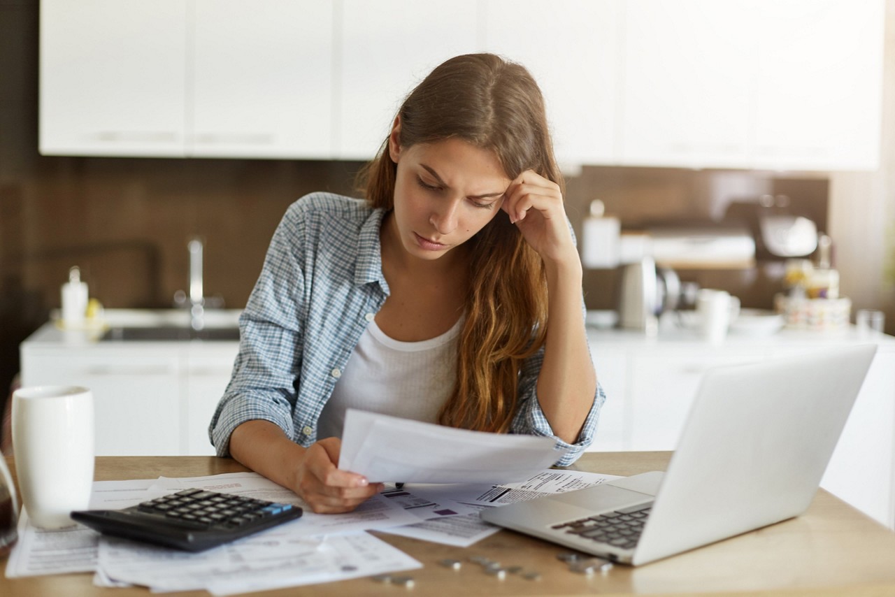 Attractive young housewife wearing shirt at home studying gas and electricity bills, checking calculations, looking at sheet of paper in her hands with serious and focused expression on her face, Attractive young housewife wearing shirt at home studying gas and electricity bills, checking calculations, looking at sheet of paper in her hands with serious and focused expression on her face