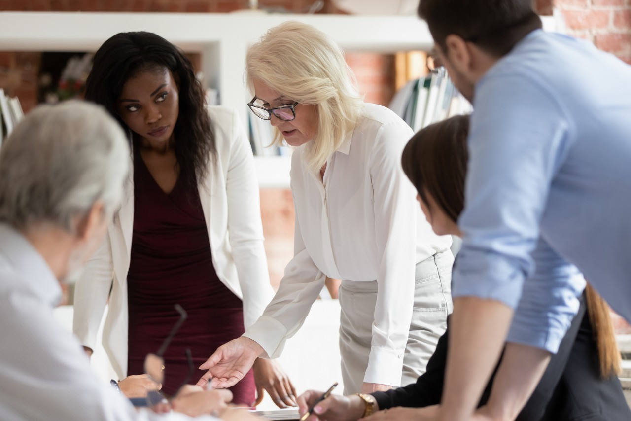 Serious mature woman supervisor teaching young staff workers explain new project plan at group meeting, focused old female teacher mentor training diverse business team at corporate office briefing