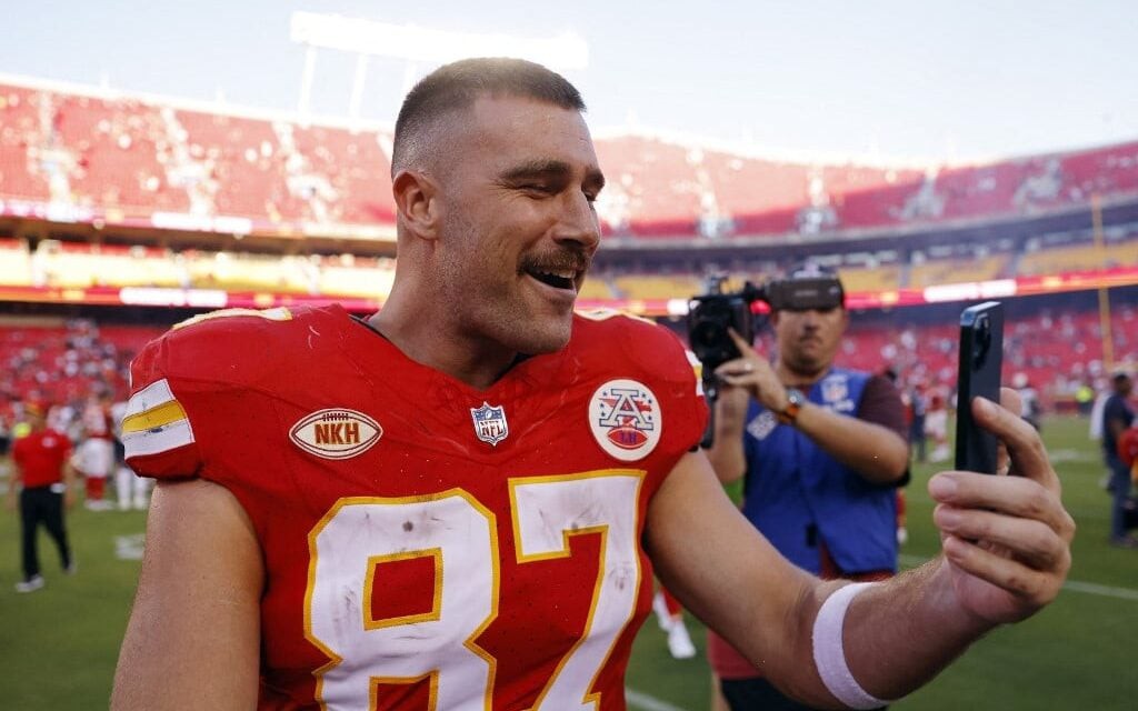 Travis Kelce, Tight End de 35 anos dos Chiefs, ainda não pensa em se aposentar da NFL. Jogador está sorrindo e segurando um celular, em video-chamada, na foto.